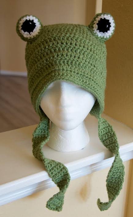 Green crocheted frog beanie with googly eyes, ear flaps and braided ties, on a mannequin head